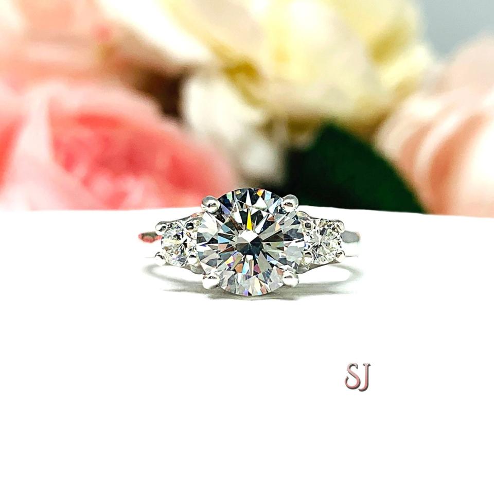 Sterling silver 3-stone engagement ring featuring an 8mm round brilliant cut near colorless cubic zirconia center stone with two smaller round CZ side stones.  Displayed on a white surface with a soft floral background.