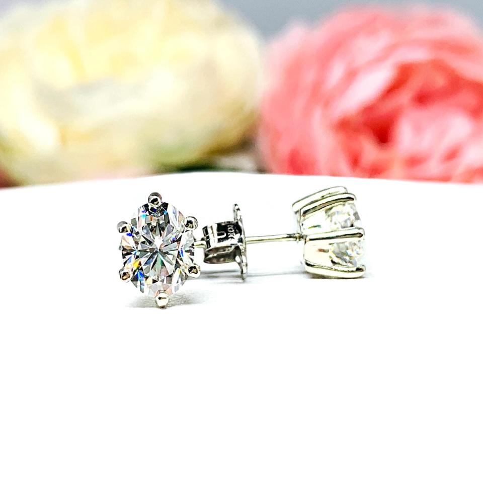 A pair of 10k white gold stud earrings set with oval brilliant cut moissanite.  The 6 prong earrings are on a white surface with the left earring facing forward to showcase the brilliant cutting of the moissanite and the right earring facing to the right to display the center post and butterfly friction clasp.