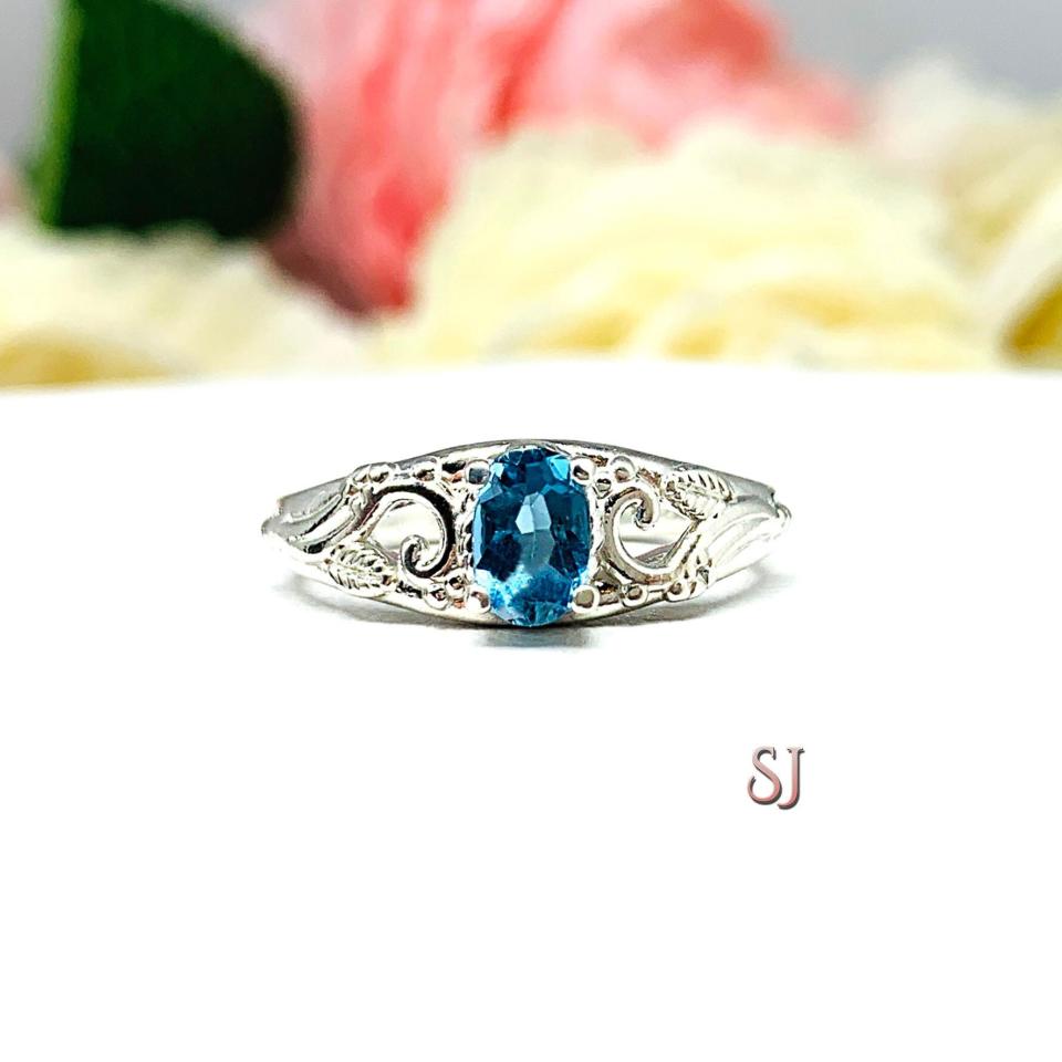 A sterling silver ring with a 6x4mm oval medium blue tone London blue topaz gemstone set in a delicate filigree design. Displayed on a white surface with a pastel floral background. The letters 'SJ' appear in the bottom right corner.