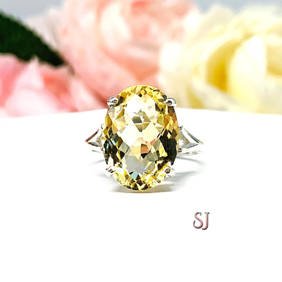 A sterling silver ring featuring a stunning, 16x12mm oval cut, golden citrine gemstone. Displayed on a white surface with soft pastel floral background.