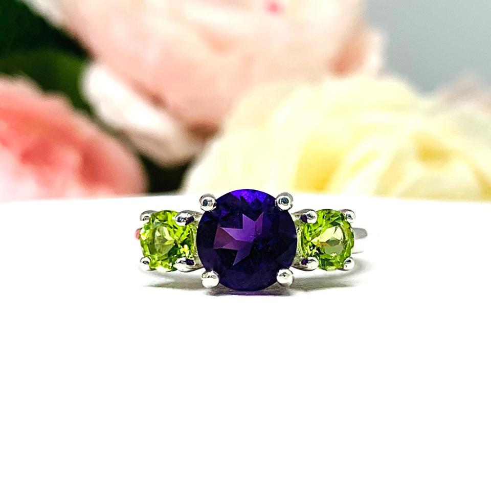 Three-stone sterling silver ring featuring a natural 8mm round African amethyst flanked by two 5mm round Arizona peridot gemstones. The rich purple and bright green hues contrast beautifully against the soft pink and white floral backdrop, emphasizing the clarity and vibrancy of the stones. A timeless setting that blends bold elegance with earthy charm.