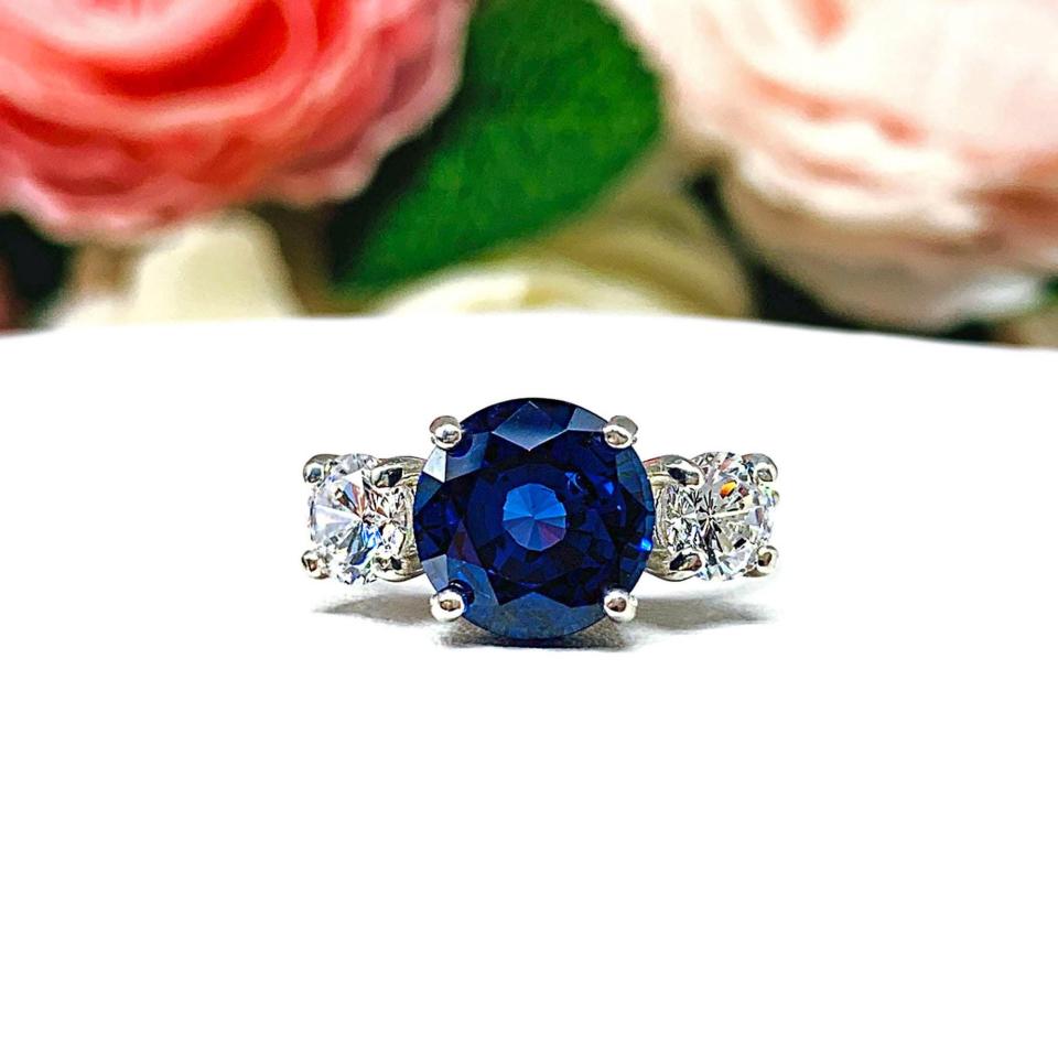 Sterling silver 3-stone ring with a round lab-created blue sapphire center flanked by smaller round brilliant cut cubic zirconias on each side. The ring is set against a white surface with a background of pink and white flowers.