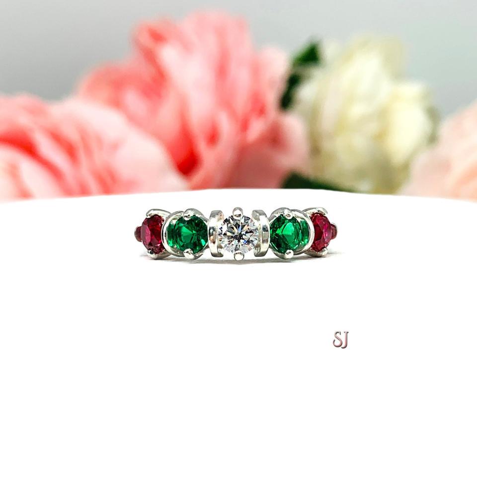 A sterling silver ring with alternating 4mm round lab ruby and emeralds, with a central round cz representing a "Christmas star." The ring resting on a white surface with blurred pink and white flowers in the background.