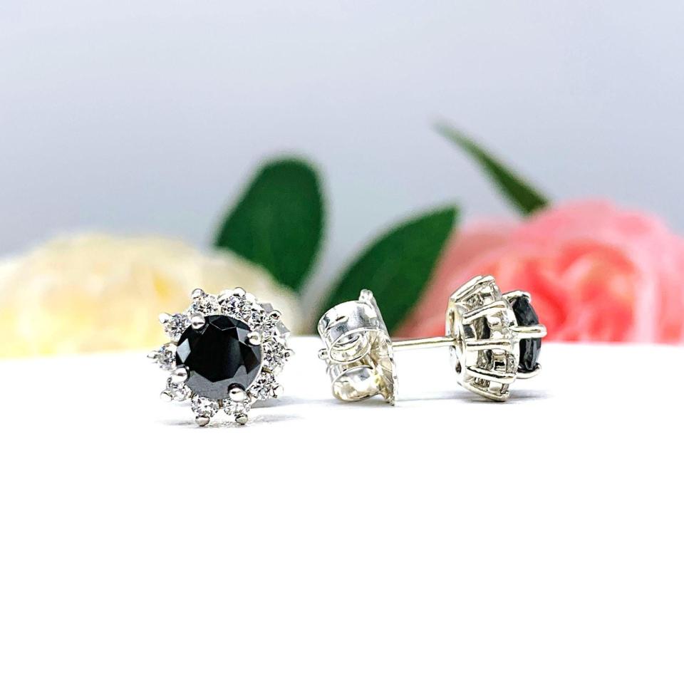 A pair of sterling silver stud earrings set with round black moissanites with several small colorless moissanites surrounding them in a halo. The earrings are shown on a white surface with a background of soft-focus pink and white flowers.