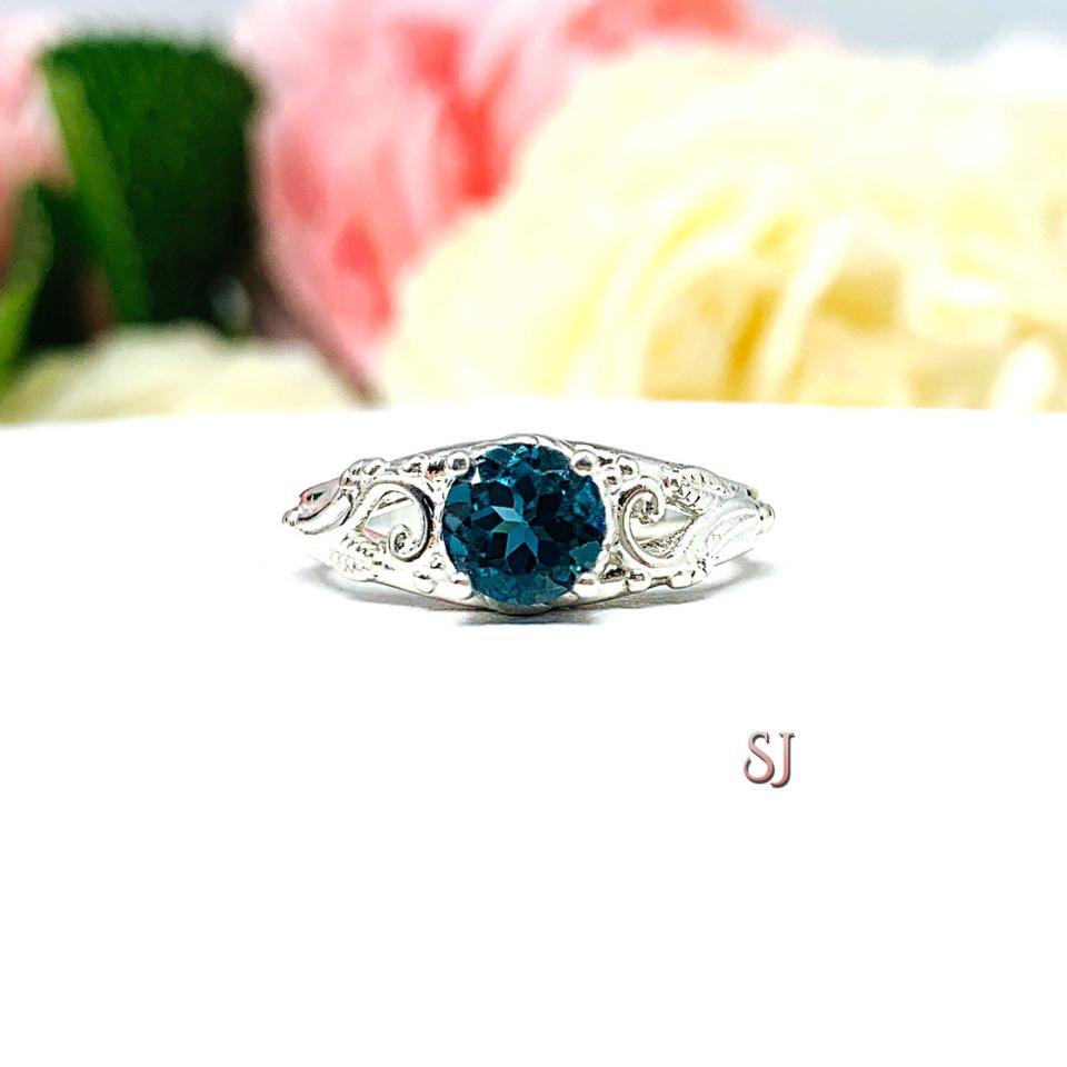 A sterling silver ring with a richly saturated 6mm round London blue topaz gemstone set in a delicate filigree design. Displayed on a white surface with a pastel floral background. The letters 'SJ' appear in the bottom right corner.