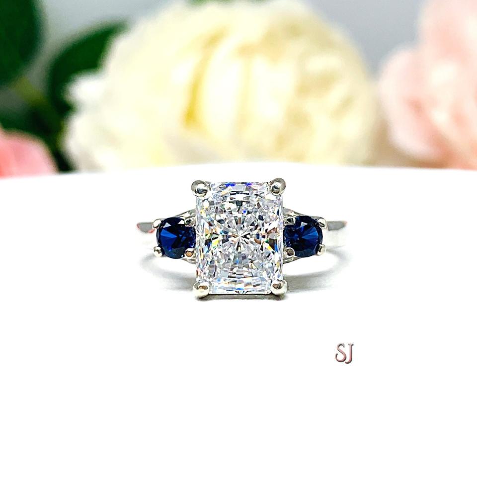 A sterling silver 3-stone ring featuring a 9x7mm radiant cut cz flanked by two 3.5mm round lab blue sapphires. Displayed on a white surface with a pastel floral background.