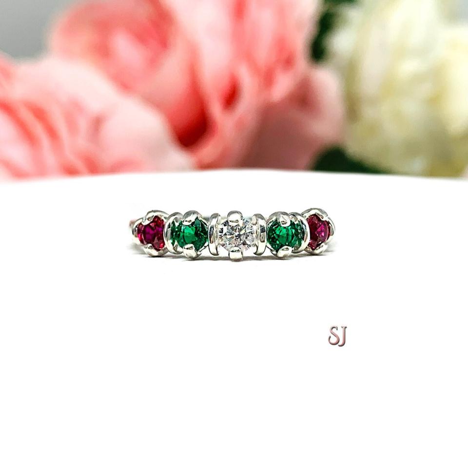 A sterling silver ring with alternating 3mm round lab ruby and emeralds, with a central round cz representing a "Christmas star." The ring resting on a white surface with blurred pink and white flowers in the background.