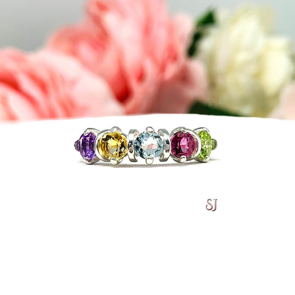 A sterling silver "spring season" ring with five gemstones. The 4mm round gemstones are purple (amethyst), yellow (citrine), blue (sky blue topaz), pink (pink topaz), and green (peridot). Displayed on a white surface with pastel floral background.