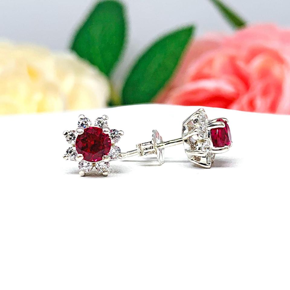 A pair of silver stud earrings set with round lab rubies with several small czs surrounding them in a halo. The earrings are shown on a white background with the left earring facing forward showcasing the mixed brilliant faceting and deep red color of the rubies and clear clarity of the czs, and the right earring facing to the right displaying the center post and butterfly friction clasp.