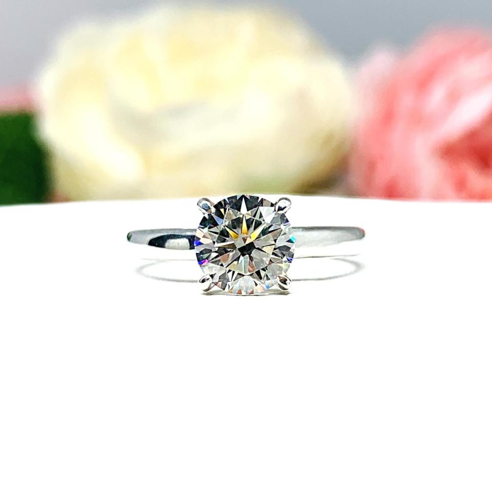 Close-up view of a near colorless (G/H color) round brilliant cut cubic zirconia solitaire ring in a claw prong cathedral setting. The ring is made of sterling silver and features a sleek, minimalist design. The setting is simple yet elegant, allowing the center stone to take center stage. The ring is displayed against a soft, blurred background of pastel-colored flowers, creating a romantic and delicate ambiance.