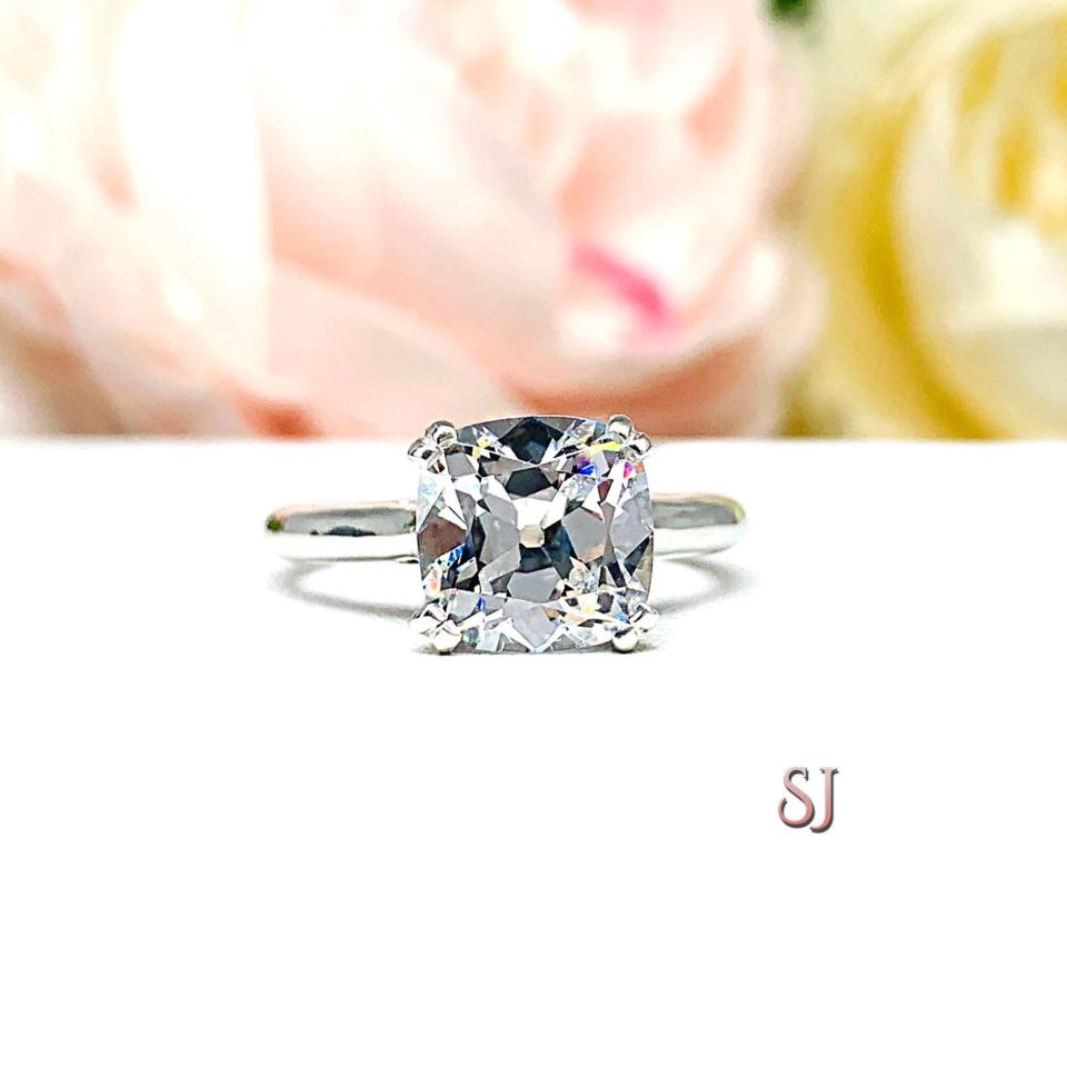 A sterling silver split-prong cathedral engagement ring set with a colorless old mine cut cubic zirconia. The ring is set against a white background with a blurred image of pink and white flowers in the background.