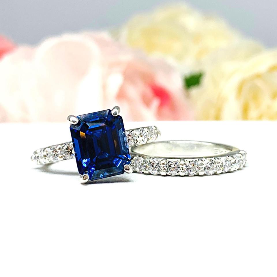 Close-up view of a stunning sterling silver emerald-cut blue sapphire engagement ring and a matching wedding band. The engagement ring features a vibrant dark blue sapphire with pave cubic zirconia side stones. The wedding band is a half-eternity band, also in sterling silver, with a continuous row of smaller white cubic zirconias. The rings are displayed against a blurred background of soft pink and white roses.