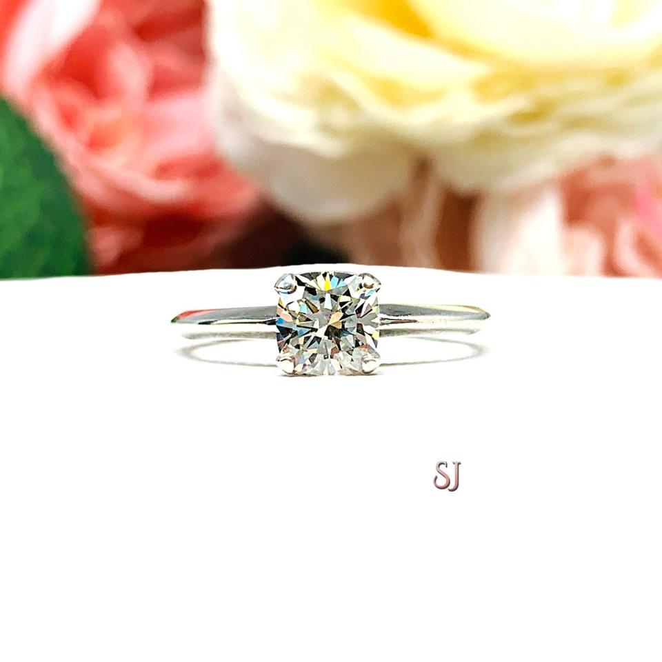 Sterling silver engagement ring set with an 6mm brilliant cushion near colorless cubic zirconia. The ring is resting on a white surface with a background of soft-focus pink and white flowers.
