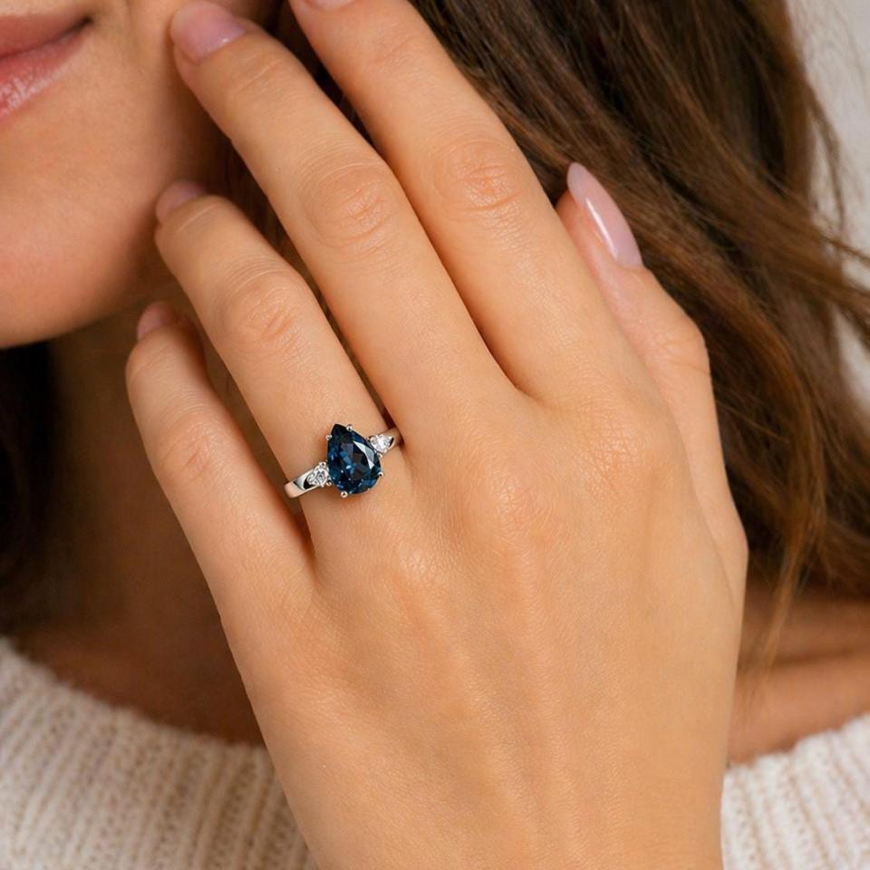 Sterling silver ring, featuring a London blue topaz flanked by two round moissanites, shown worn on the hand of a woman. Model image for size and styling reference — not a photo of the actual ring.