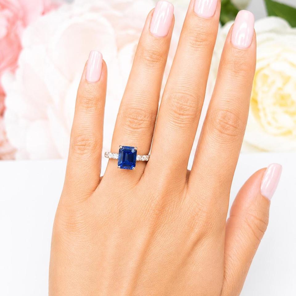 A woman's hand wearing a sterling silver engagement ring featuring a 10x8mm emerald cut blue sapphire and 2.5mm round czs on the band.  Model image for size and styling reference — not a photo of the actual ring.
