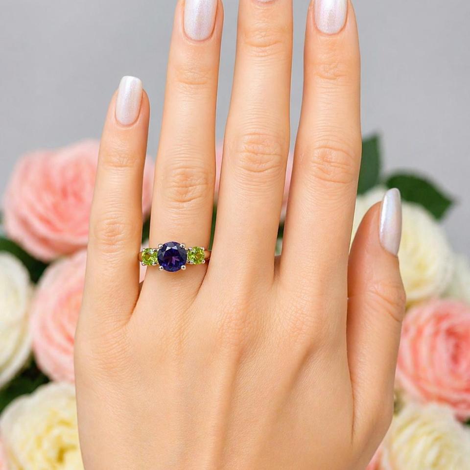 A woman's hand wearing a three-stone sterling silver ring featuring a natural 8mm round African amethyst flanked by two 5mm round Arizona peridot gemstones.
