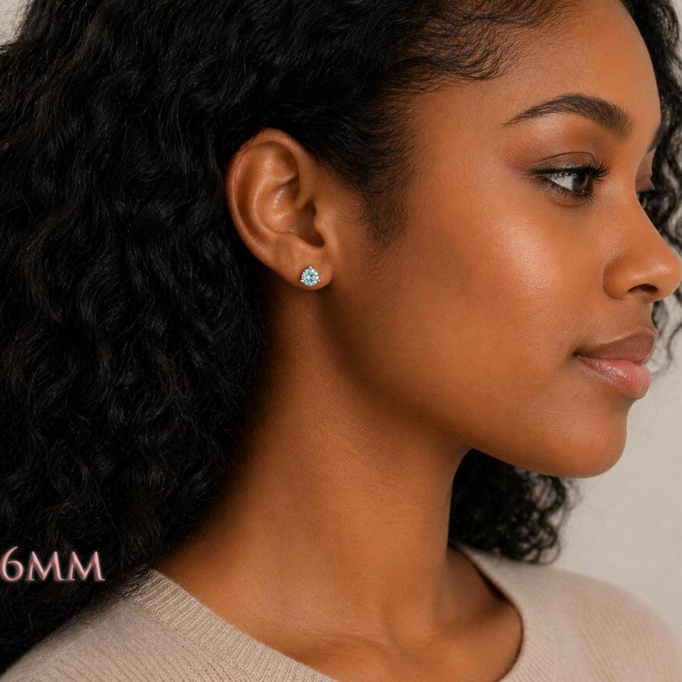 Woman wearing sterling silver stud earrings featuring 6mm round authentic blue moissanite stones in basket martini-style settings. Model image for size and styling reference — not a photo of the actual earrings.