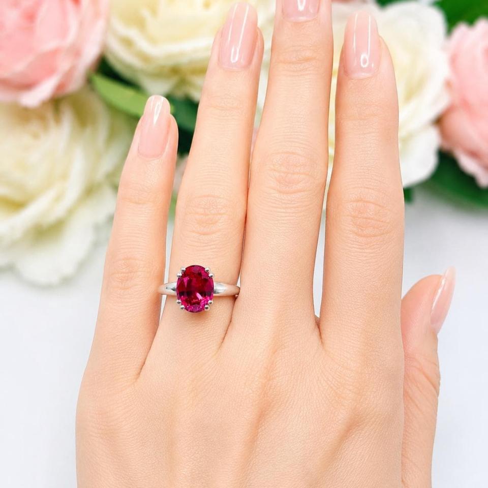 A woman's hand wearing a sterling silver ring with an oval lab created deep red ruby set in a double prong setting. Model image for size and styling reference — not a photo of the actual ring.