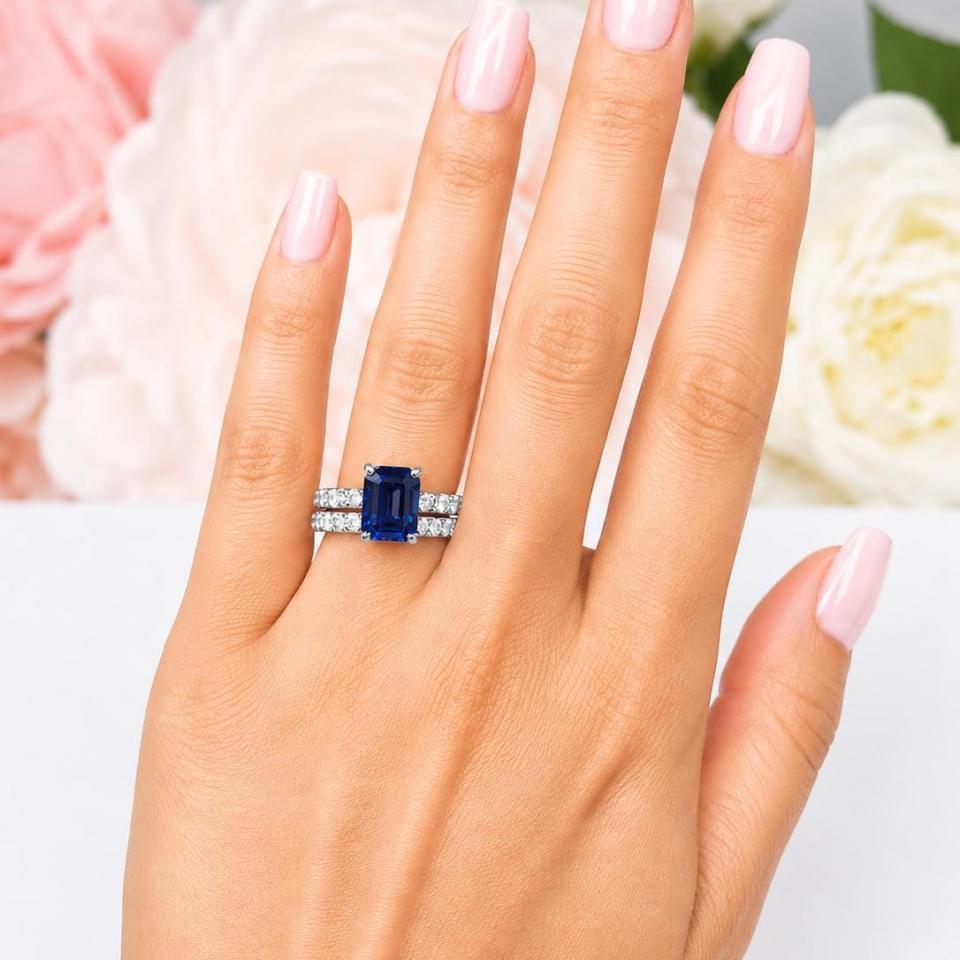 A woman's hand wearing a sterling silver engagement ring featuring a 10x8mm emerald cut blue sapphire and 2.5mm round czs on the band. Shown with matching wedding band. Model image for size and styling reference — not a photo of the actual ring.