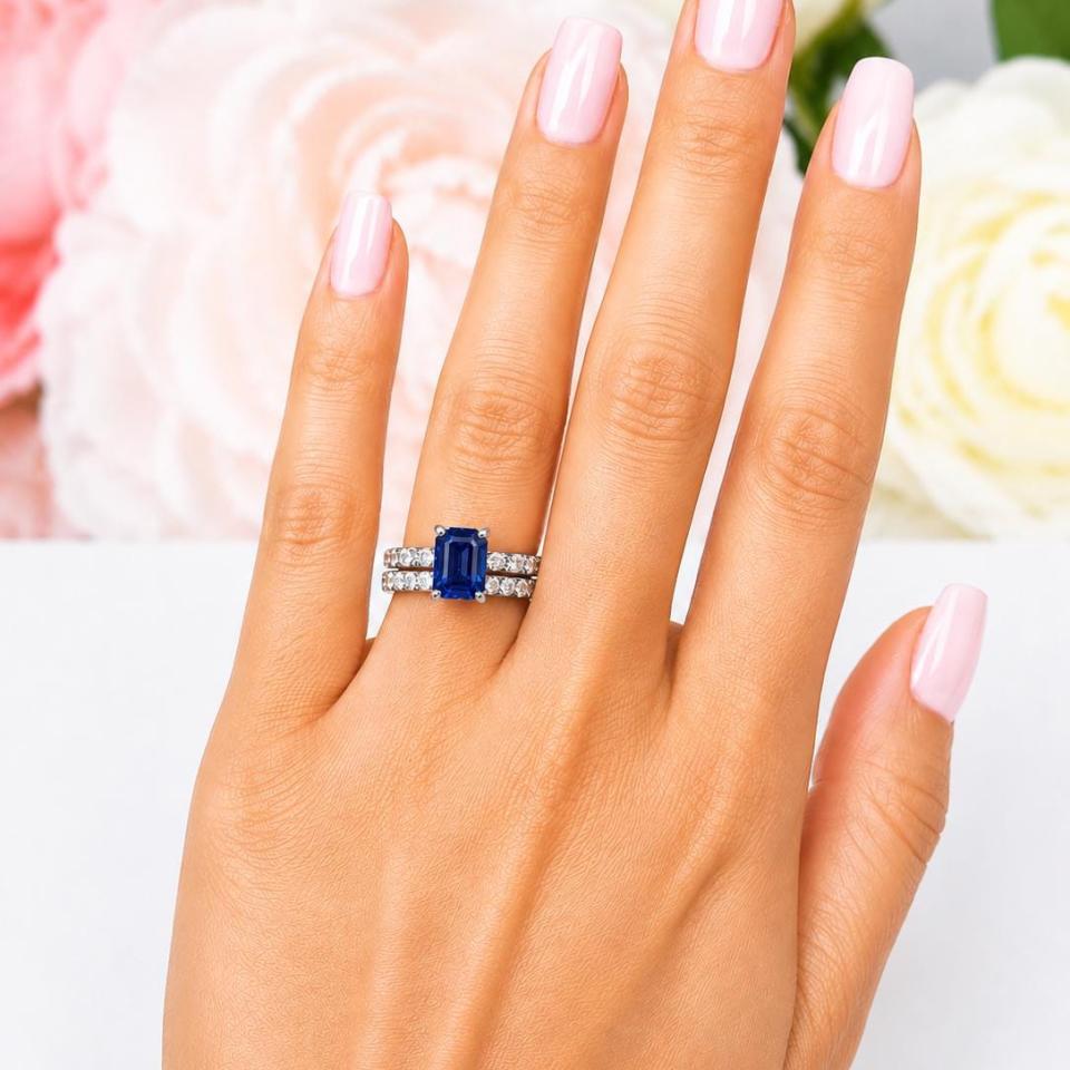 A woman's hand wearing a sterling silver engagement ring featuring an 8x6mm emerald cut blue sapphire and 2.5mm round czs on the band. Shown with matching wedding band. Model image for size and styling reference — not a photo of the actual ring.