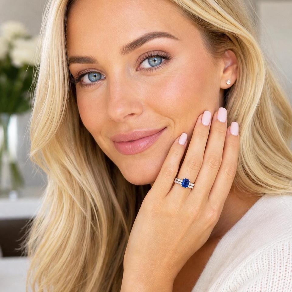 A woman wearing a sterling silver engagement ring featuring an emerald cut blue sapphire and 2.5mm round czs on the band. Shown with matching wedding band. Model image for size and styling reference — not a photo of the actual ring.