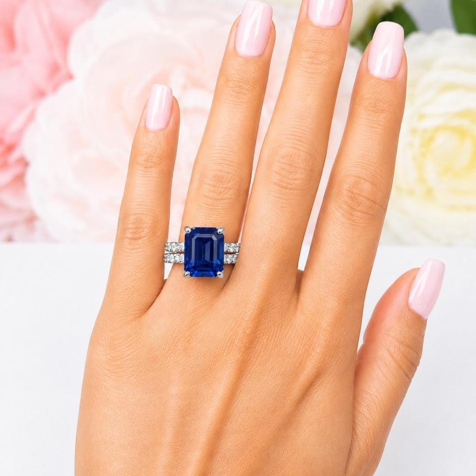 A woman's hand wearing a sterling silver engagement ring featuring a 12x10mm emerald cut blue sapphire and 2.5mm round czs on the band. Shown with matching wedding band. Model image for size and styling reference — not a photo of the actual ring.