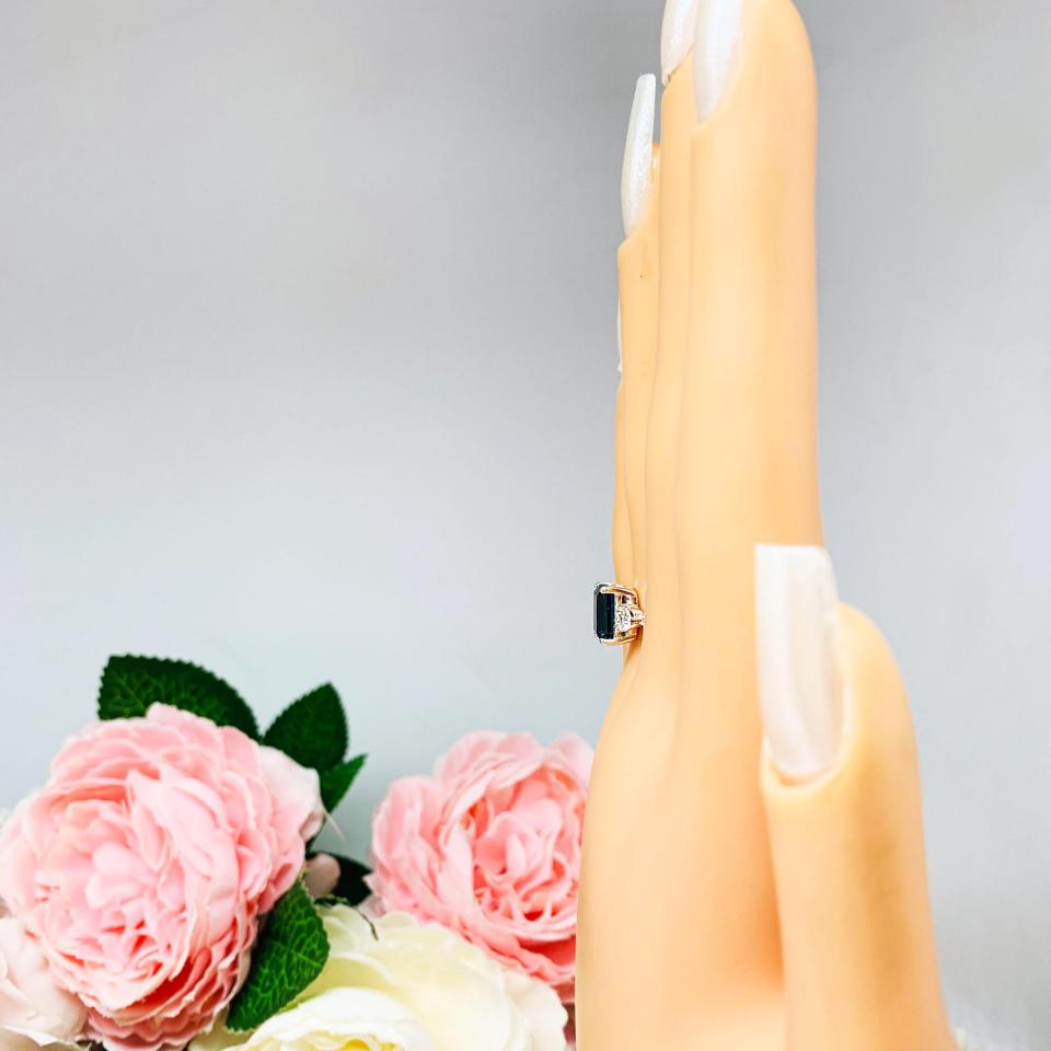 Mannequin hand with long white shimmery nails, displayed against a soft white-gray background with pink and white floral accents. The hand is wearing an 8x6mm emerald-cut London Blue Topaz ring with moissanite side stones, set in a sterling silver twist band with milgrain detailing.