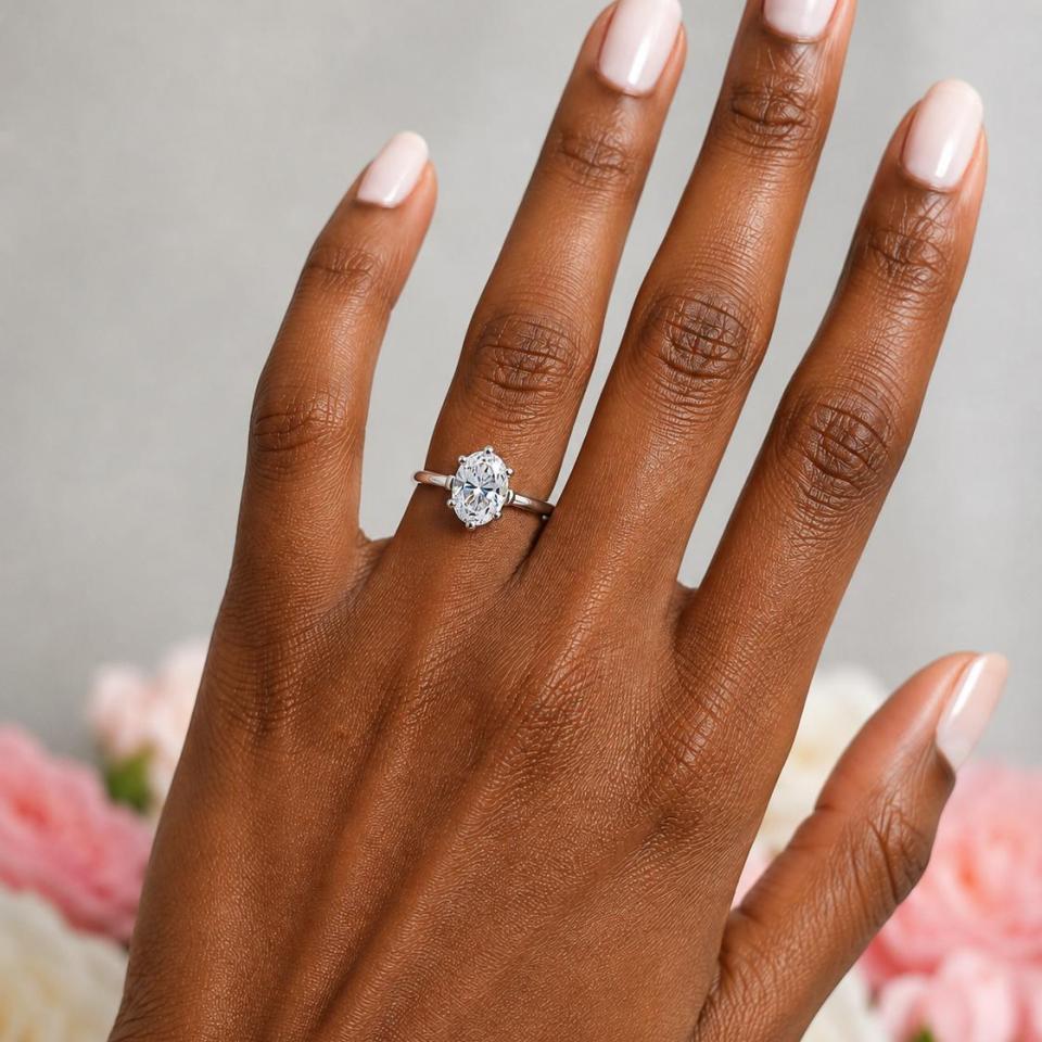 A woman's hand wearing a sterling silver 6-prong solitaire engagement ring set with a 9x7mm brilliant oval cubic zirconia. The ring features a tulip design and cathedral shank. Model image for size and styling reference — not a photo of the actual ring.