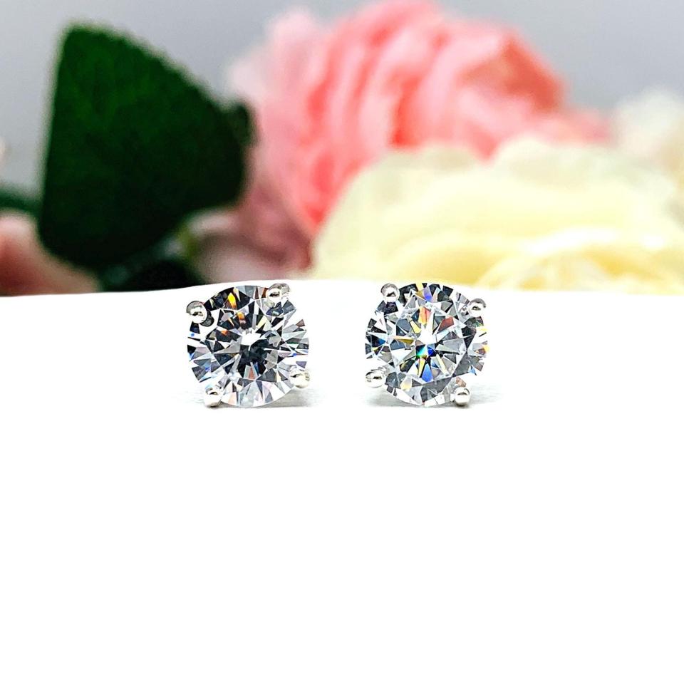 A pair of silver stud earrings set with round brilliant cut cubic zirconias.  The four prong basket earrings are on a white surface with both earrings facing forward t to showcase the brilliant cutting of the czs, center post and butterfly friction clasps.