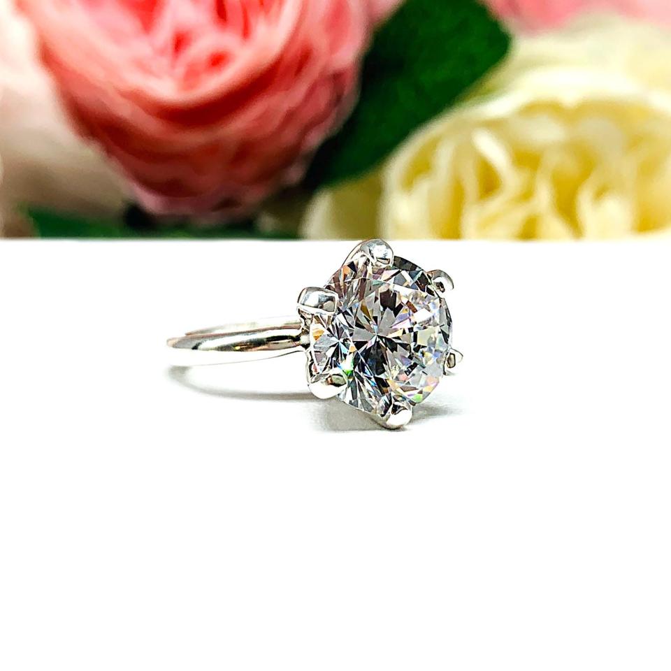 Close-up view of a 10mm near colorless (G/H color) round brilliant cut cubic zirconia solitaire engagement ring in a classic 6-prong setting. The setting is simple yet elegant, allowing the center stone to take center stage. The ring is displayed against a soft, blurred background of pastel-colored flowers, creating a romantic and delicate ambiance.