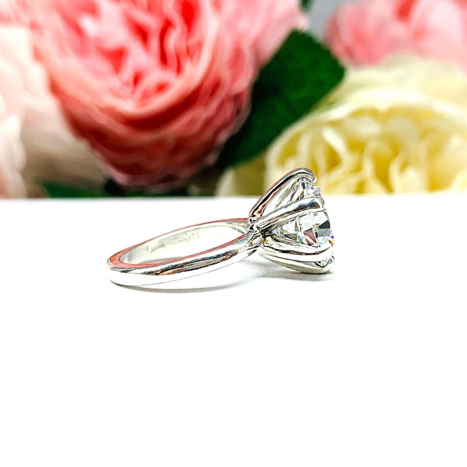 Close-up view of a 10mm near colorless (G/H color) round brilliant cut cubic zirconia solitaire engagement ring in a classic 6-prong setting. The setting is simple yet elegant, allowing the center stone to take center stage. The ring is displayed against a soft, blurred background of pastel-colored flowers, creating a romantic and delicate ambiance.