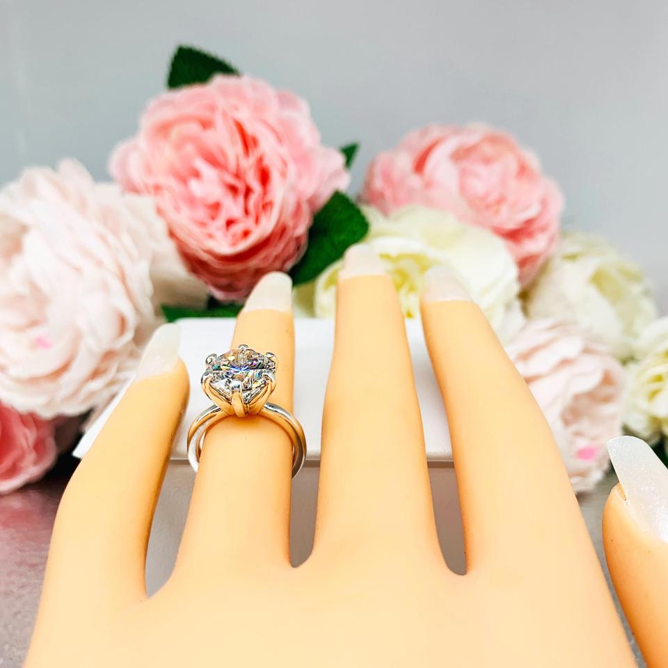 Mannequin hand with long white shimmery nails, displayed against a soft white-gray background with pink and white floral accents. The hand is wearing a 10mm near colorless (G/H color) round brilliant cut cubic zirconia solitaire engagement ring in a classic 6-prong setting. The setting is simple yet elegant, allowing the center stone to take center stage.