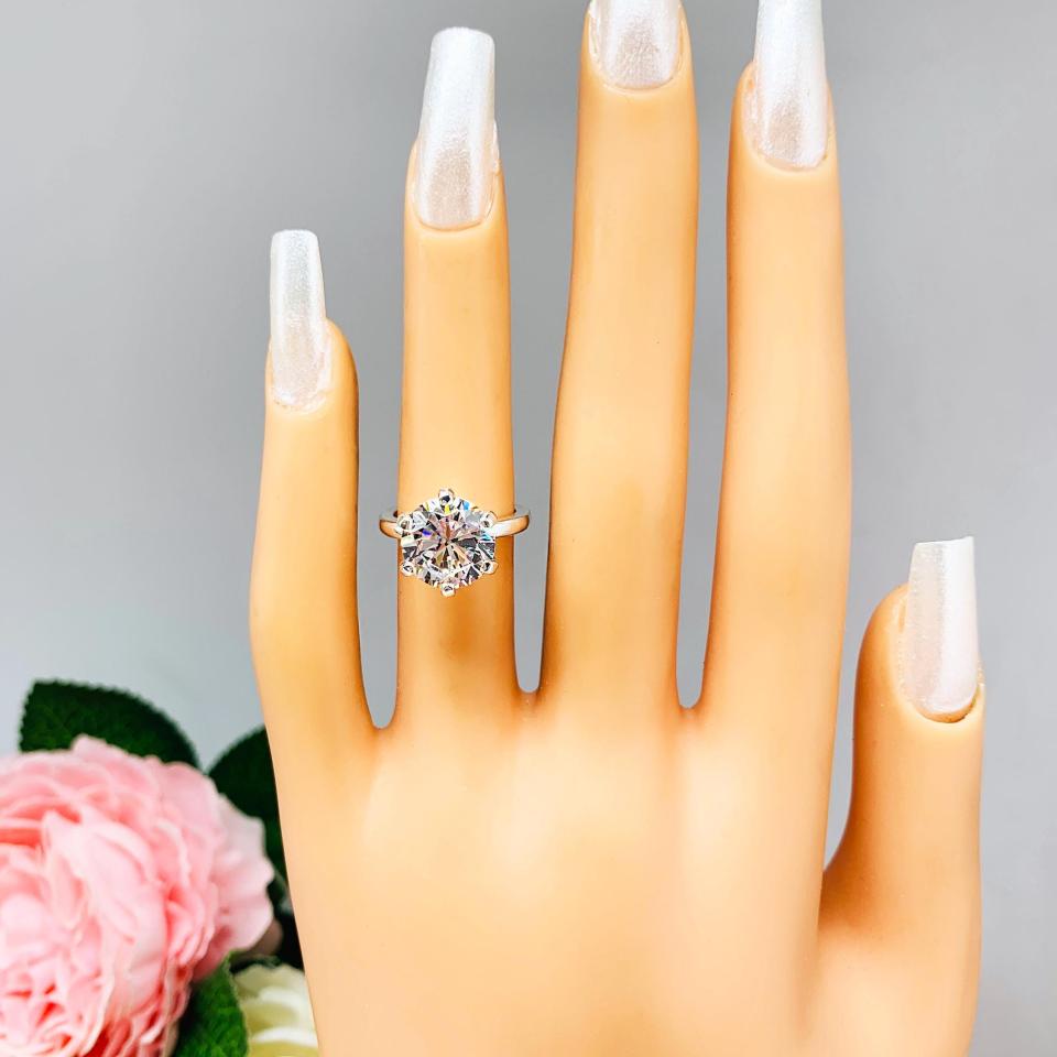 Mannequin hand with long white shimmery nails, displayed against a soft white-gray background with pink and white floral accents. The hand is wearing a 10mm near colorless (G/H color) round brilliant cut cubic zirconia solitaire engagement ring in a classic 6-prong setting. The setting is simple yet elegant, allowing the center stone to take center stage.