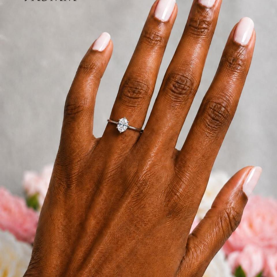 A woman's hand wearing a sterling silver 6-prong solitaire engagement ring set with a 7x5mm brilliant oval cubic zirconia. The ring features a tulip design and cathedral shank. Model image for size and styling reference — not a photo of the actual ring.