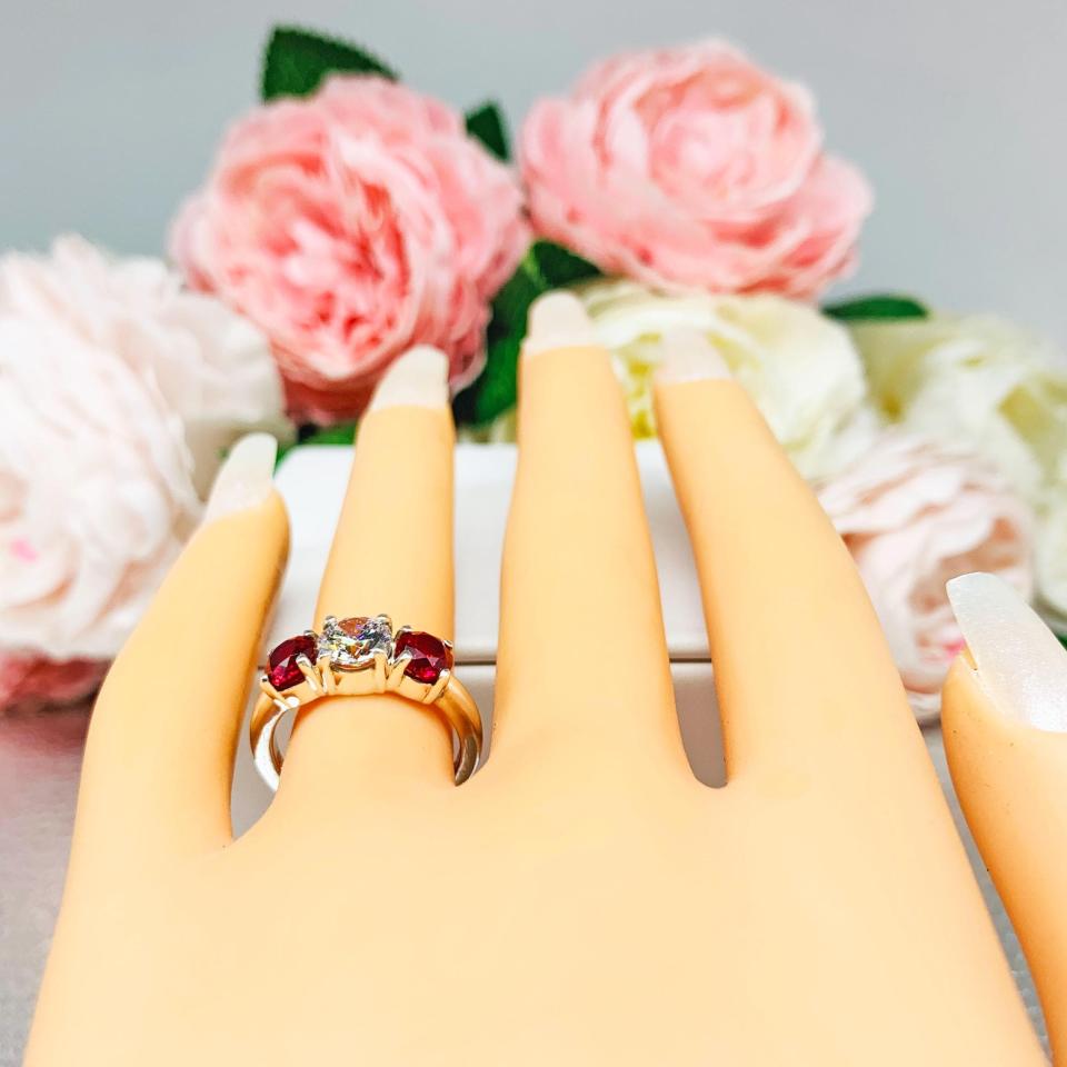 Mannequin hand with long white shimmery nails, displayed against a soft white-gray background with pink and white floral accents. The hand is wearing a sterling silver 3-stone ring with a 6mm round brilliant cut cubic zirconia center that radiates icy sparkle, flanked by two 5mm round deep red lab-created rubies.
