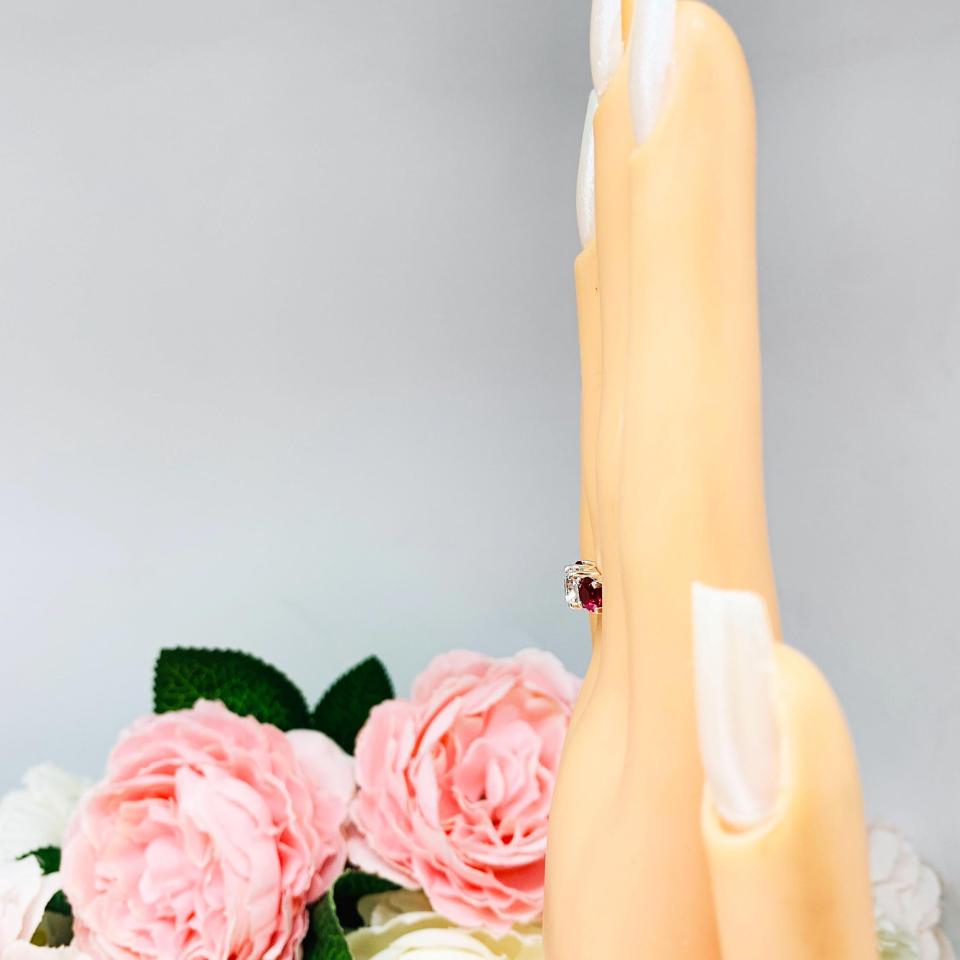 Mannequin hand with long white shimmery nails, displayed against a soft white-gray background with pink and white floral accents. The hand is wearing a sterling silver 3-stone ring with a 6mm round brilliant cut cubic zirconia center that radiates icy sparkle, flanked by two 5mm round deep red lab-created rubies.