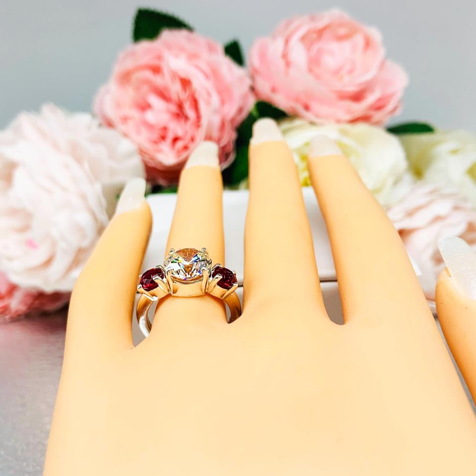 Mannequin hand with long white shimmery nails, displayed against a soft white-gray background with pink and white floral accents. The hand is wearing a sterling silver 3-stone ring with a 9mm near colorless round brilliant cut cubic zirconia center that radiates icy sparkle, flanked by two 5mm round deep red lab-created rubies.