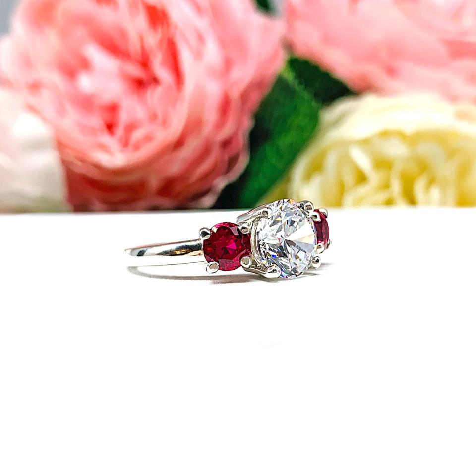 Sterling silver 3-stone ring with a round brilliant cut colorless (D color) cubic zirconia center flanked by two deep red lab-created rubies. The ring is set on a white surface with a background of pink and white flowers.