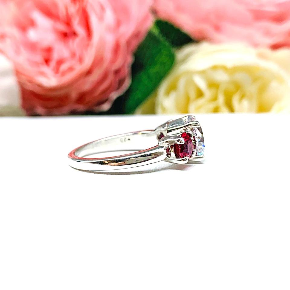 Sterling silver 3-stone ring with a round brilliant cut colorless (D color) cubic zirconia center flanked by two deep red lab-created rubies. The ring is set on a white surface with a background of pink and white flowers.