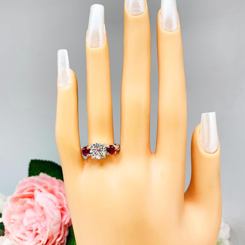 Mannequin hand with long white shimmery nails, displayed against a soft white-gray background with pink and white floral accents. The hand is wearing a sterling silver 3-stone ring with an 8mm round brilliant cut cubic zirconia center that radiates icy sparkle, flanked by two 5mm round deep red lab-created rubies.