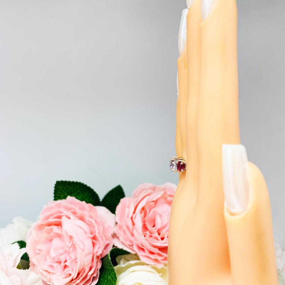 Mannequin hand with long white shimmery nails, displayed against a soft white-gray background with pink and white floral accents. The hand is wearing a sterling silver 3-stone ring with a 9mm near colorless round brilliant cut cubic zirconia center that radiates icy sparkle, flanked by two 5mm round deep red lab-created rubies.