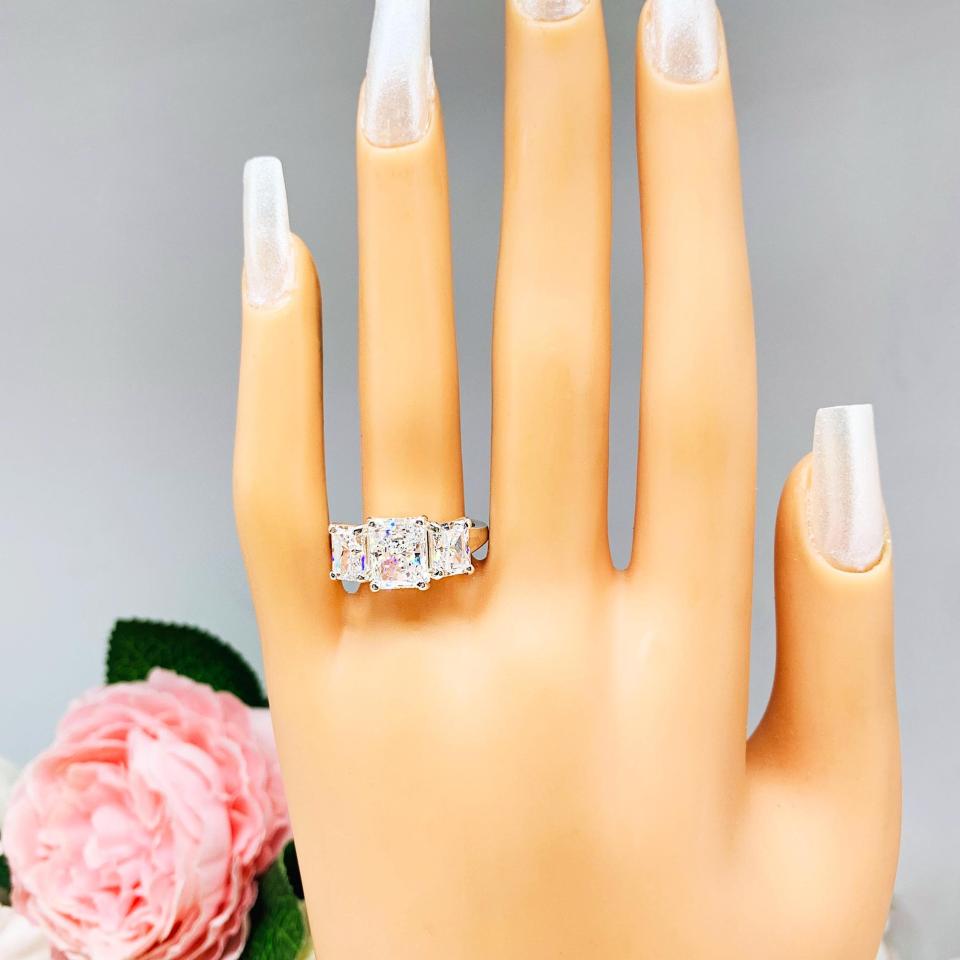 Mannequin hand with long white shimmery nails, displayed against a soft white-gray background with pink and white floral accents. The hand is wearing a sterling silver 3-stone engagement ring featuring a 9x7mm radiant cut cubic zirconia center stone with two 7x5mm radiant CZ side stones. The highly polished wide band tapers for comfort. Displayed on a white surface with soft floral background.