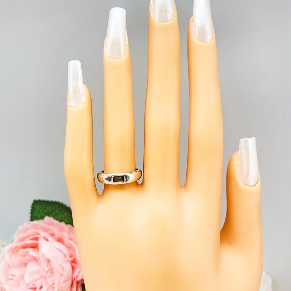 Mannequin hand with long white shimmery nails, displayed against a soft white-gray background with pink and white floral accents. The hand is wearing a sterling silver band featuring a 5×3 mm emerald‑cut green tourmaline set flush in a polished band.