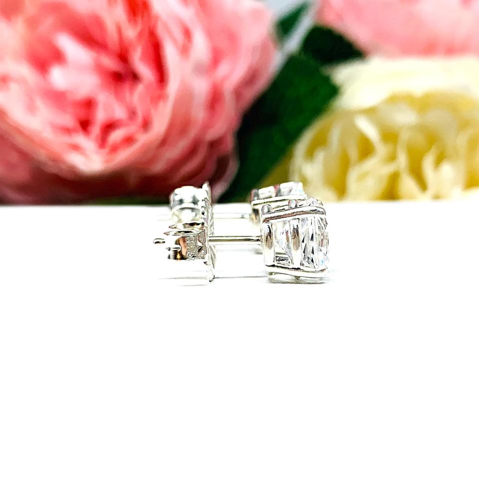 A pair of sterling silver basket stud earrings set with round mixed-brilliant cut lab white sapphires shown on a white surface with pink and white flowers in the background.