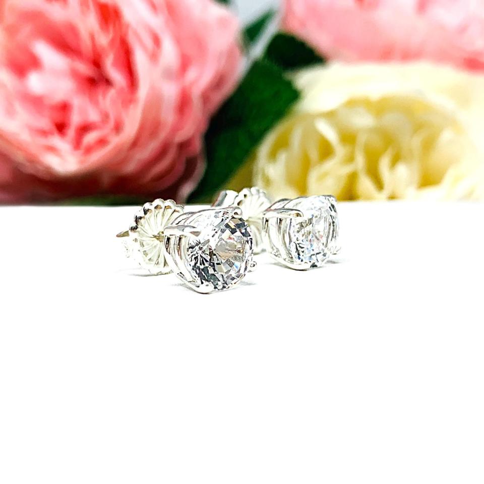 A pair of sterling silver basket stud earrings set with round mixed-brilliant cut lab white sapphires shown on a white surface with pink and white flowers in the background.