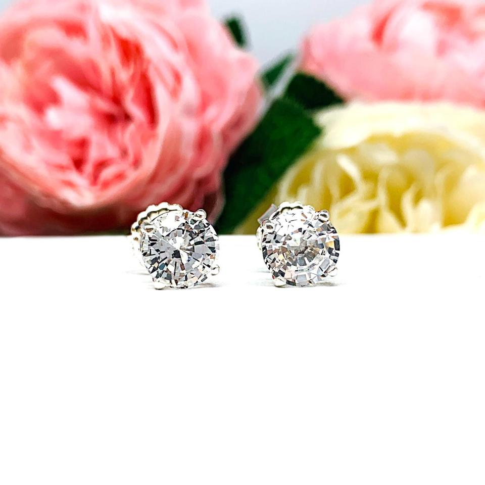 A pair of sterling silver basket stud earrings set with round mixed-brilliant cut lab white sapphires shown on a white surface with pink and white flowers in the background.