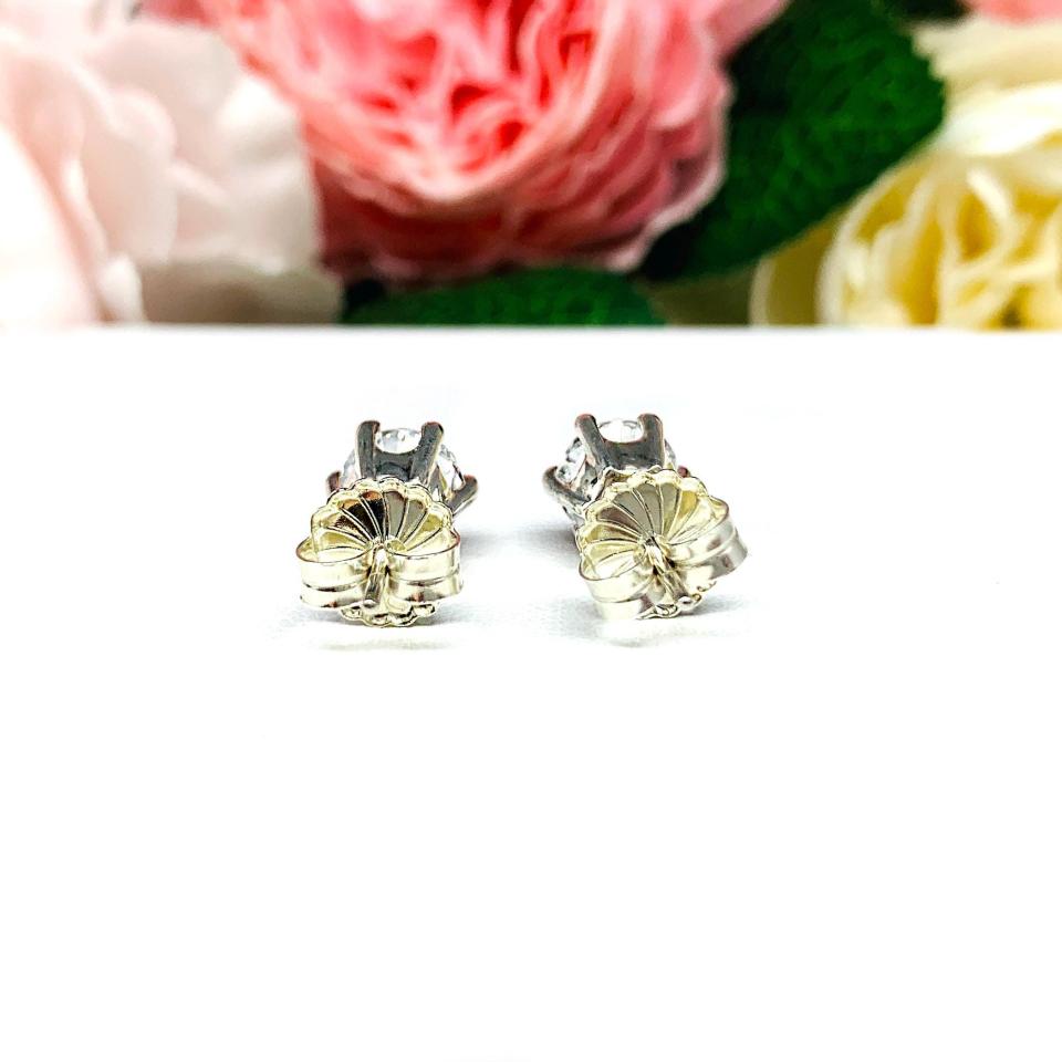 A pair of sterling silver six prong stud earrings set with round brilliant lab white sapphires. The earrings have center posts and butterfly clasps and are displayed on a white surface with a floral background.