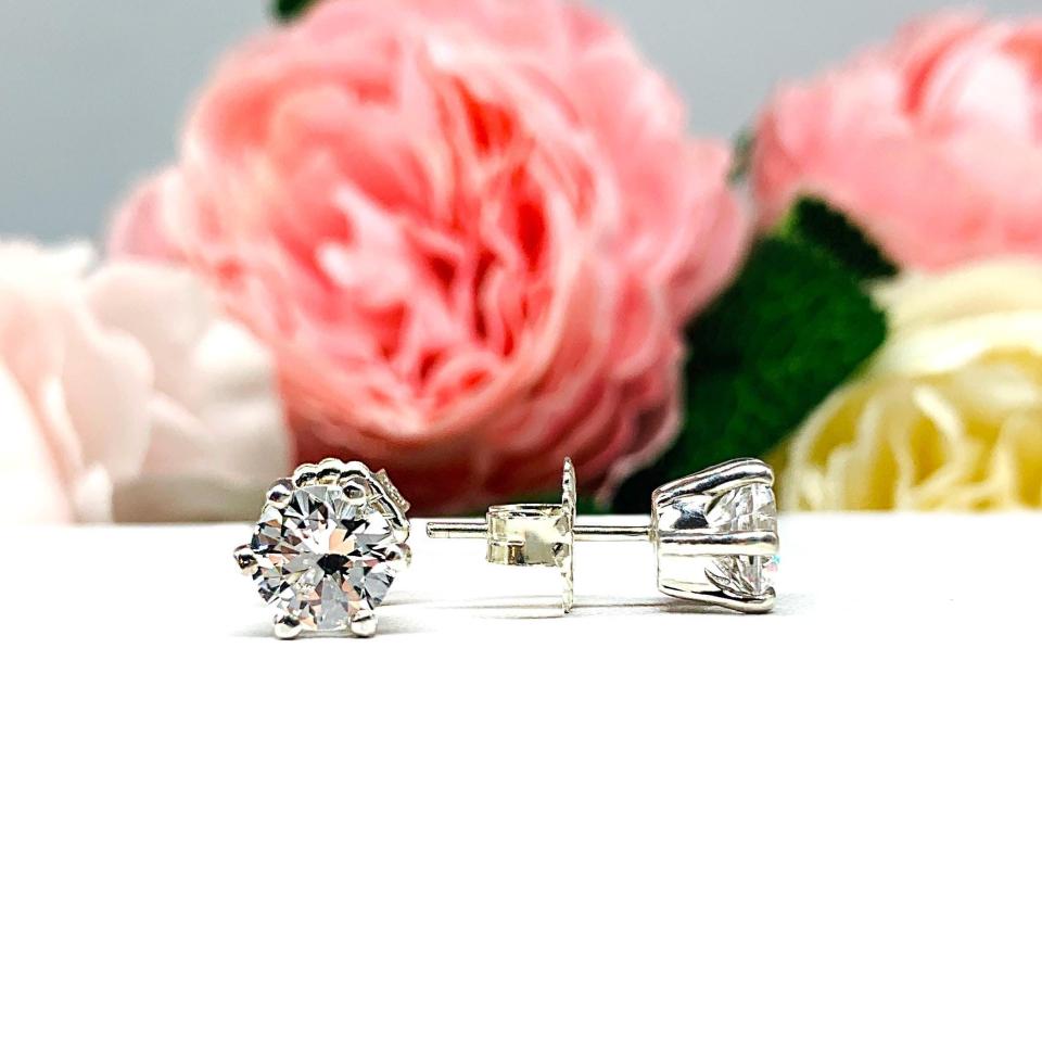 A pair of sterling silver six prong stud earrings set with round brilliant lab white sapphires. The earrings have center posts and butterfly clasps and are displayed on a white surface with a floral background.