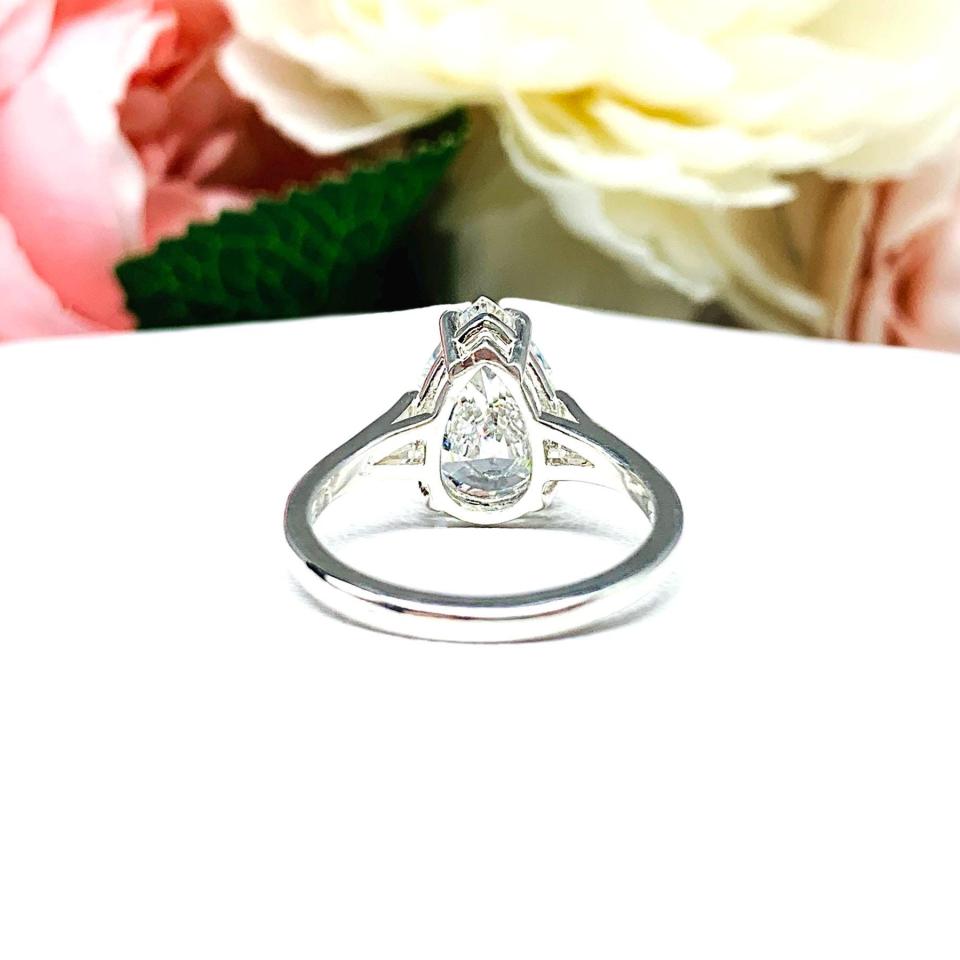 An elegant sterling silver engagement ring featuring a 13x9mm near colorless precision-cut pear cubic zirconia. Displayed on a white surface with a floral background.