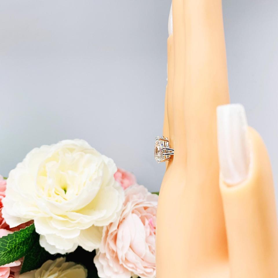 Mannequin hand with long white shimmery nails, displayed against a soft white-gray background with pink and white floral accents. The hand is wearing an elegant 4-prong sterling silver cathedral engagement ring set featuring a 13x9mm near colorless precision-cut pear cubic zirconia. Displayed on a white surface with a floral background.